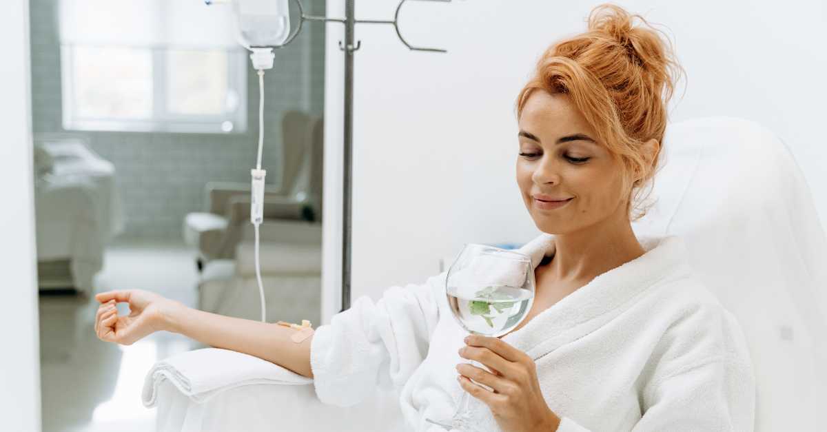 Waist up portrait view of the charming woman in white bathrobe sitting in armchair and receiving NAD+ IV infusion. She is holding glass of lemon beverage and smiling.