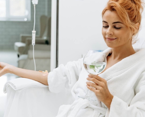 Waist up portrait view of the charming woman sitting in armchair and receiving IV therpy She is holding glass of lemon beverage and smiling.