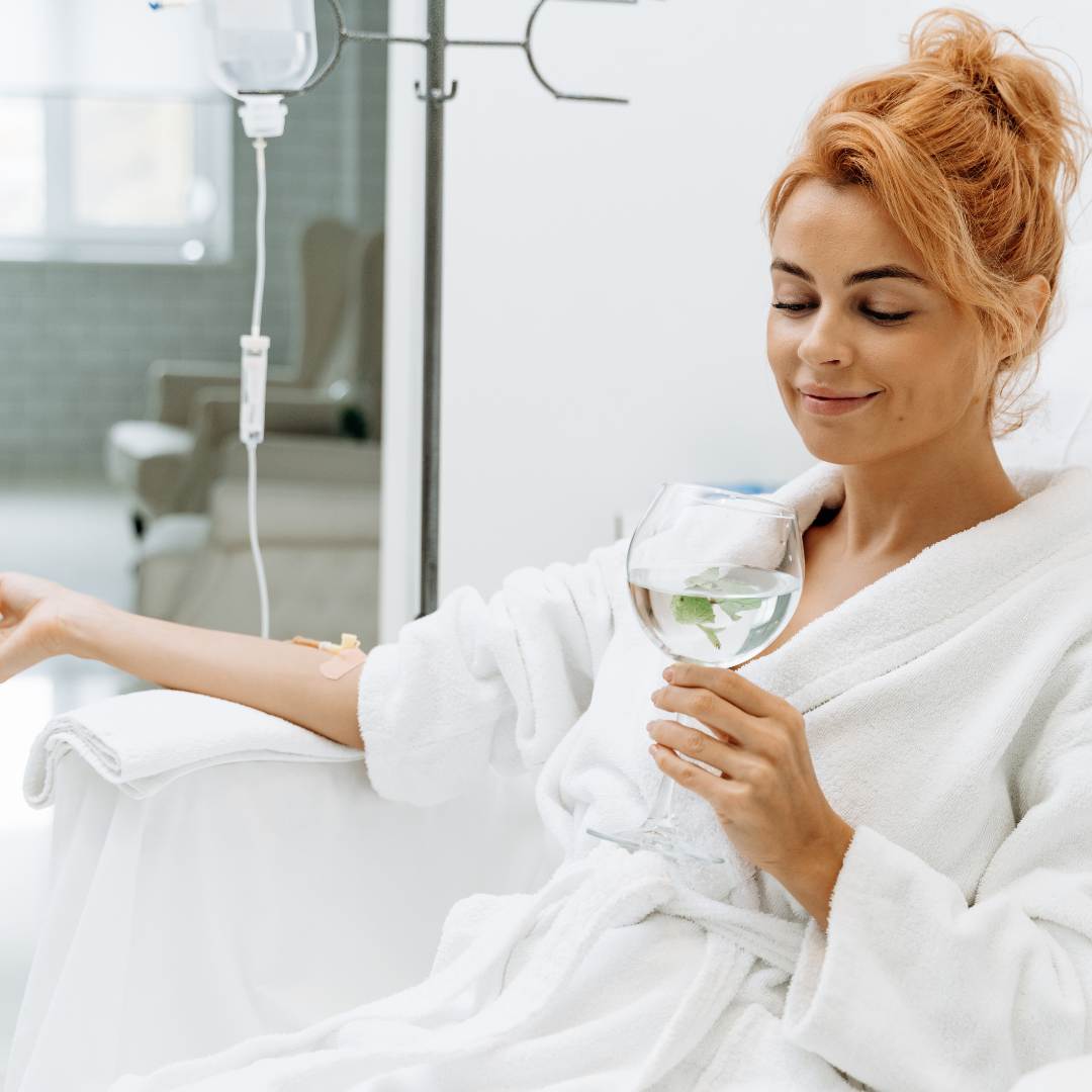 Waist up portrait view of the charming woman sitting in armchair and receiving IV therpy She is holding glass of lemon beverage and smiling.