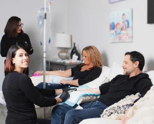 A friendly nurse with the IV Nurses administers IV hydration to a man and woman relaxing on a living room couch, illustrating the convenience of mobile IV therapy at home compared to traditional IV bars.