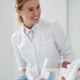 Side view portrait of charming woman in white bathrobe sitting in armchair and holding glass of lemon water. Physician in sterile gloves checking IV infusion.