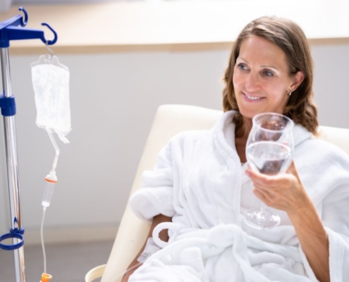 A senior woman in a white robe smiling while receiving IV vitamin therapy and holding a glass of water, illustrating wellness support for aging bodies.